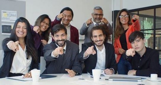 A diverse group of colleagues smiles and points at the camera