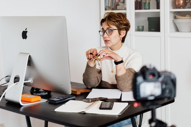 A red haired woman wearing glasses works on her computer, camera equipment for recording visible