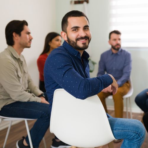 A group of employees sits in a circle discussion, while one man turn to the camera and smiles