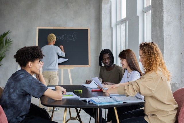 4 colleagues collaborate around a meeting room table while another writes on a chalkboard