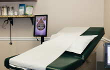 Examination table in a medical room with white paper cover and green cushioning, beside a wall shelf and sign.