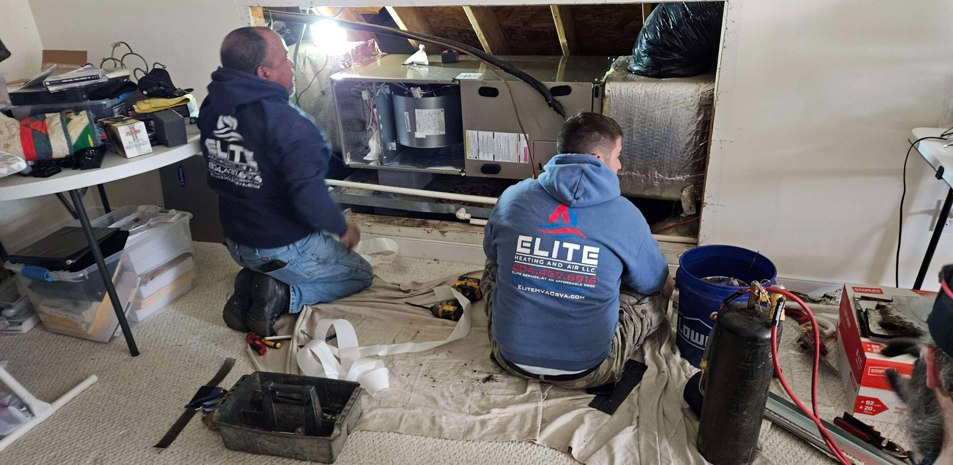 Two workers kneel beside plumbing under a sink, using tools in a cluttered utility room.