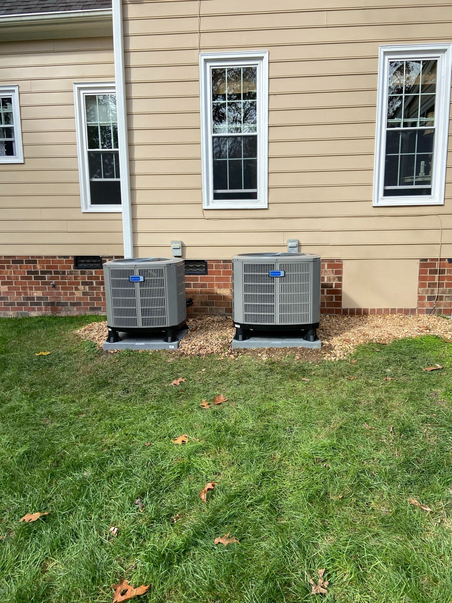 Two outdoor HVAC units beside a beige house with brick foundation and grass yard.