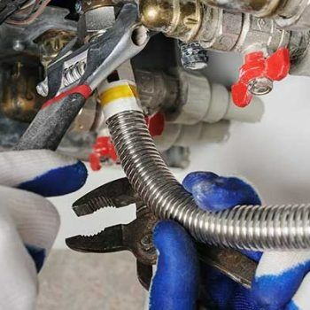 Gloved hands use pliers to tighten a corrugated metal hose on plumbing fittings with red valves
