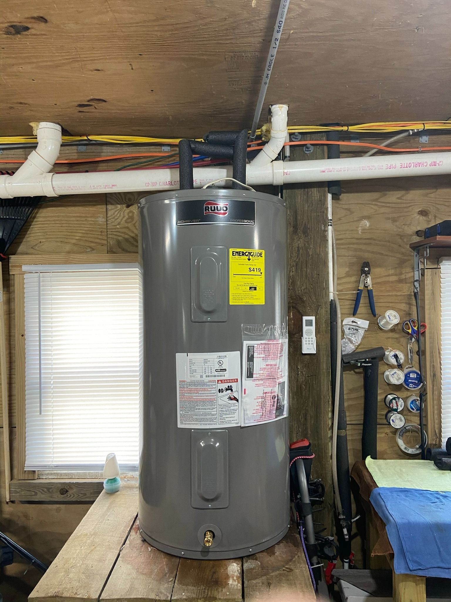 Gray water heater in a basement or utility room with exposed pipes and a window.
