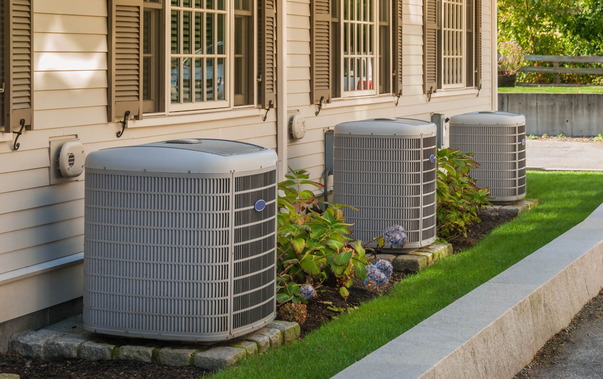 Three gray outdoor HVAC units beside a beige house with windows and a small landscaped strip