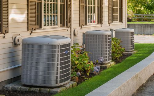 Three gray outdoor HVAC units beside a beige house with windows and a small landscaped strip