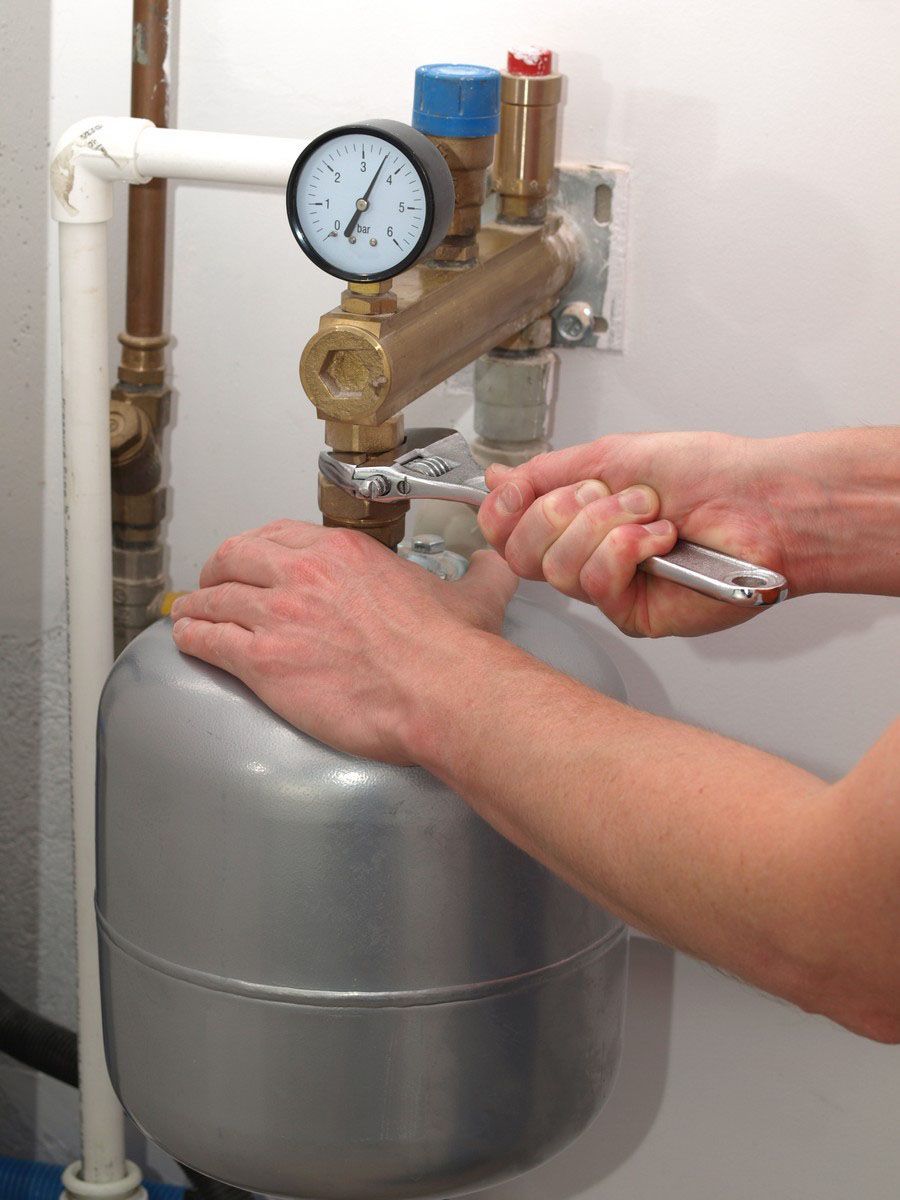 Person adjusting a boiler pressure valve beside a gauge and gray expansion tank in a utility room