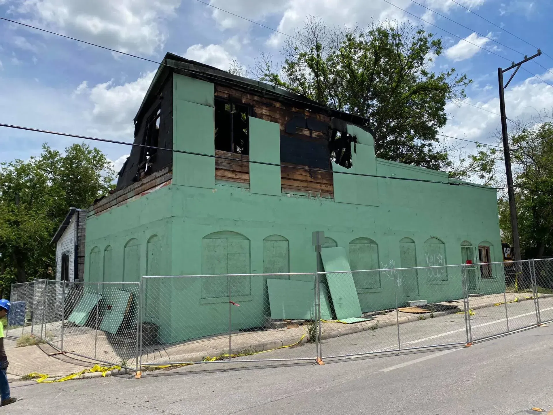 Green building with fire damage to the upper level; fenced off, on a street.