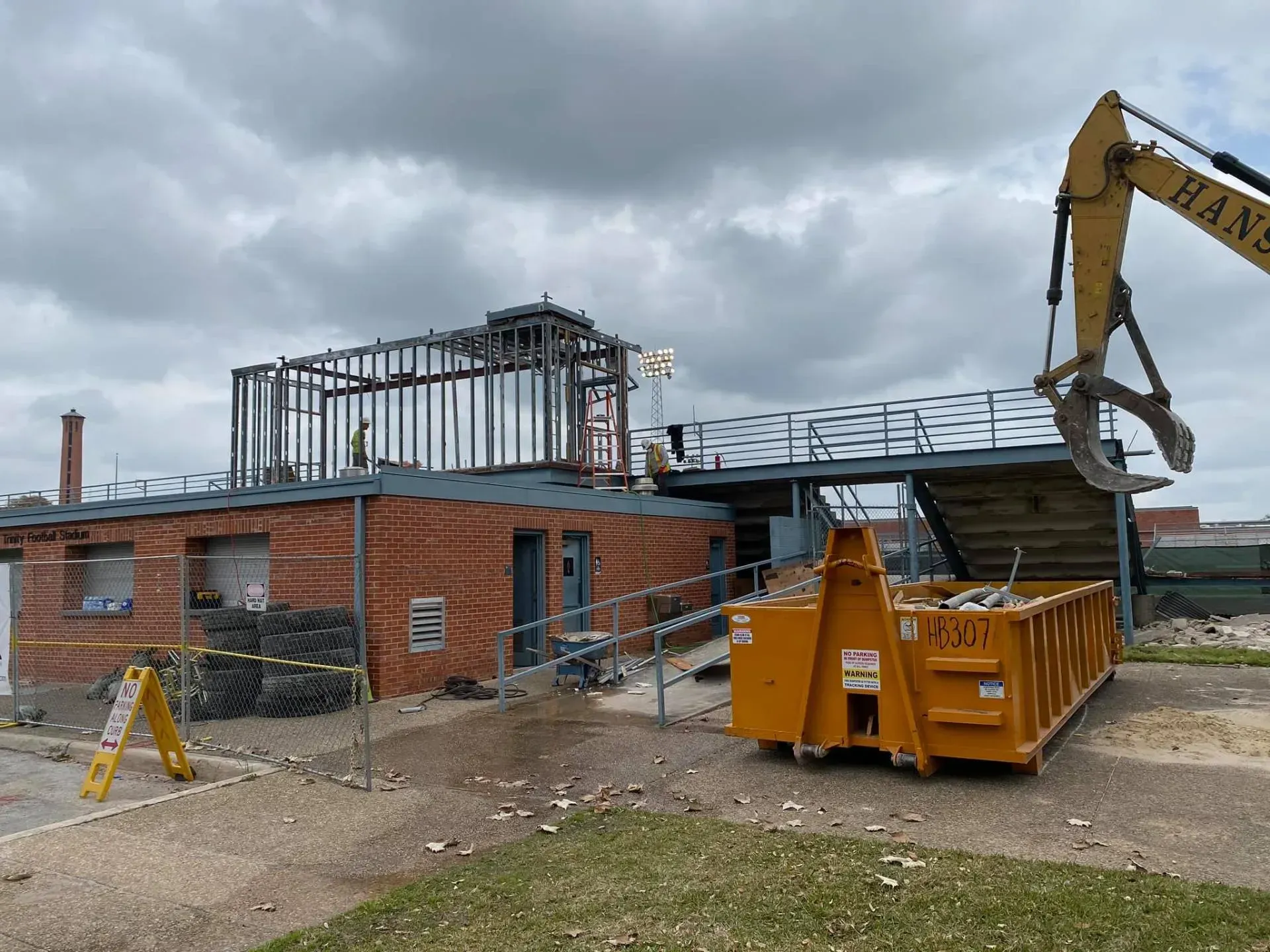 Building partially demolished by an excavator; yellow dumpster, brick exterior, cloudy sky.
