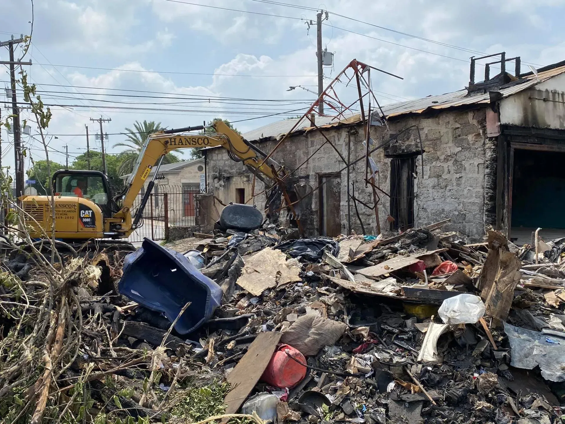 An excavator demolishes a damaged building amid debris. Blue sky, bright daylight.