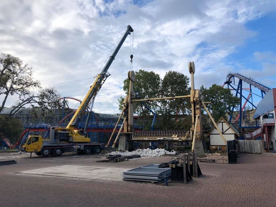 A crane dismantling a large amusement park ride with a wooden structure; other rides and buildings visible.