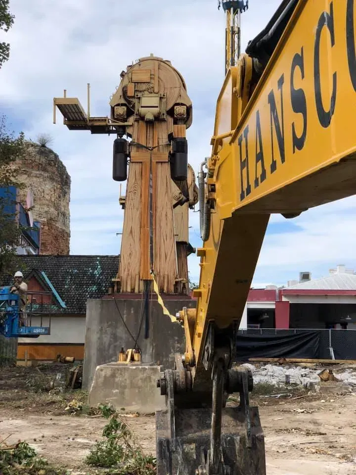 Yellow excavator arm facing a large, tan amusement ride structure. Construction site setting.