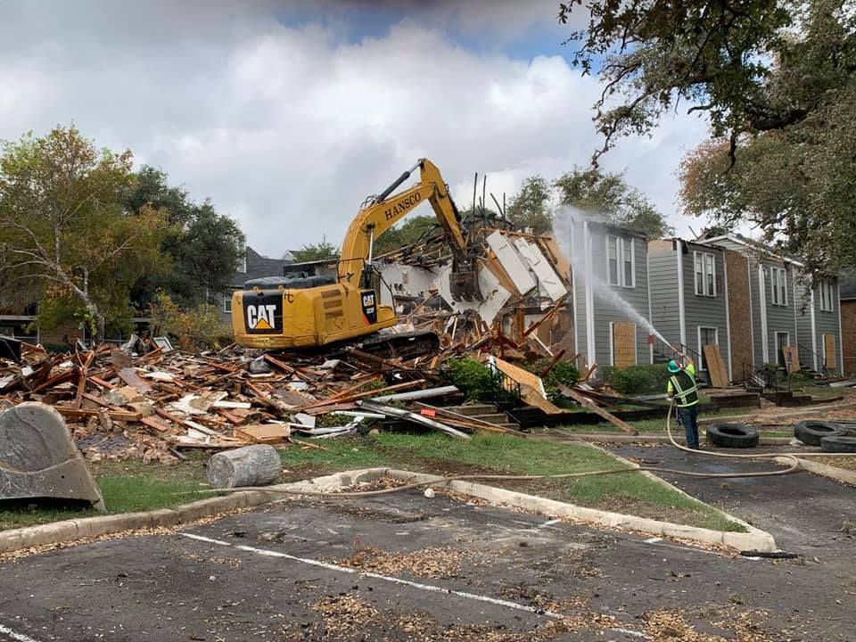 Demolition of a building by a yellow excavator with a worker spraying water to control dust.