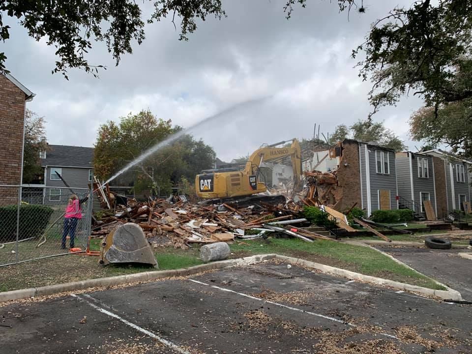 Demolition of an apartment building with an excavator spraying water. Person holding hose nearby. Cloudy sky.