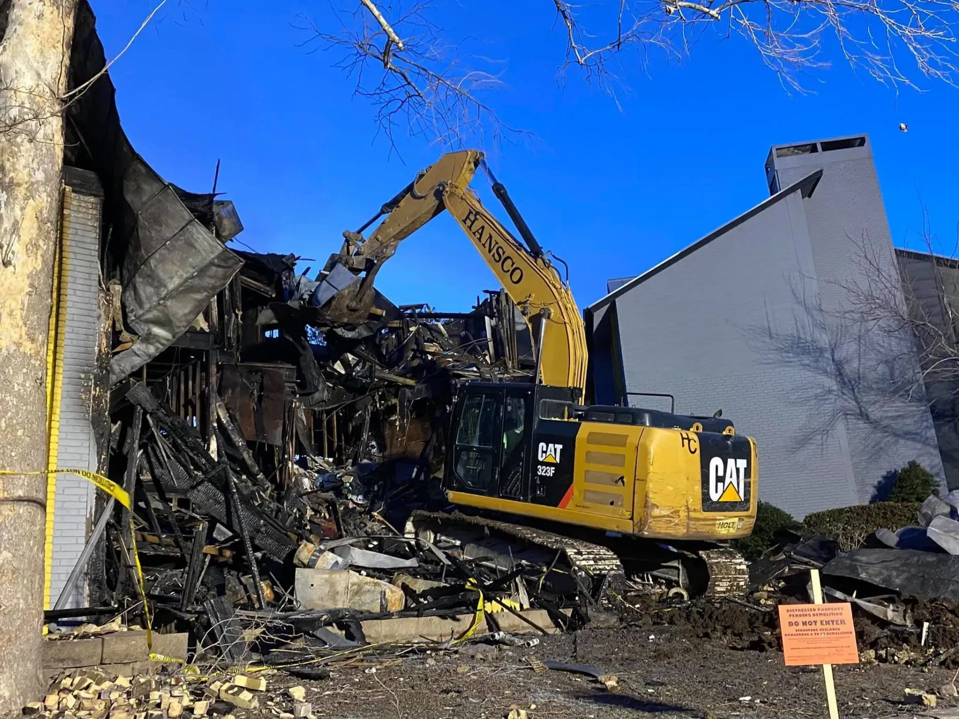 An excavator demolishes a burned building. The structure is charred black with yellow caution tape nearby.
