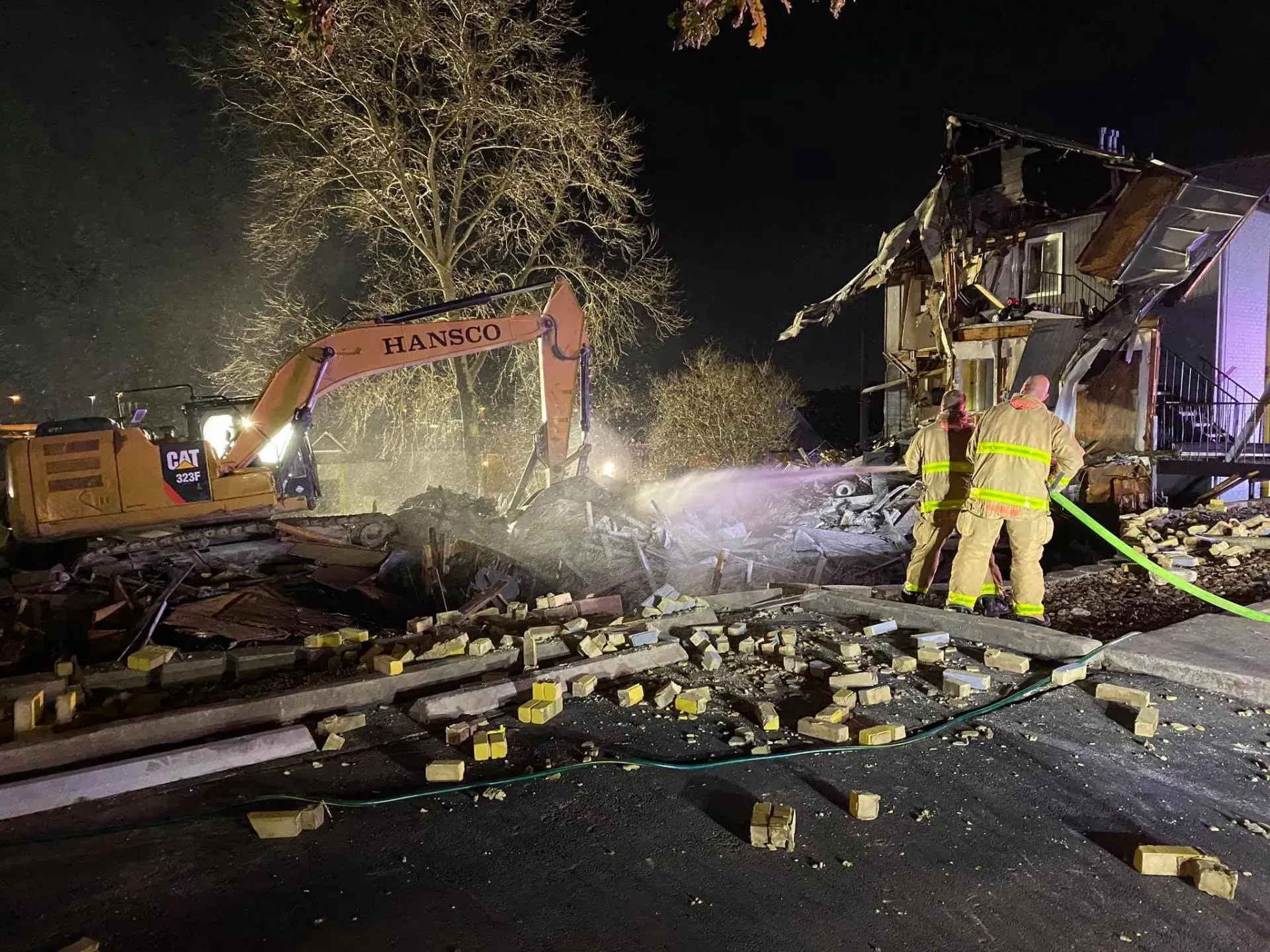 Firefighters and excavator at a burned building at night. Debris and water on the ground.
