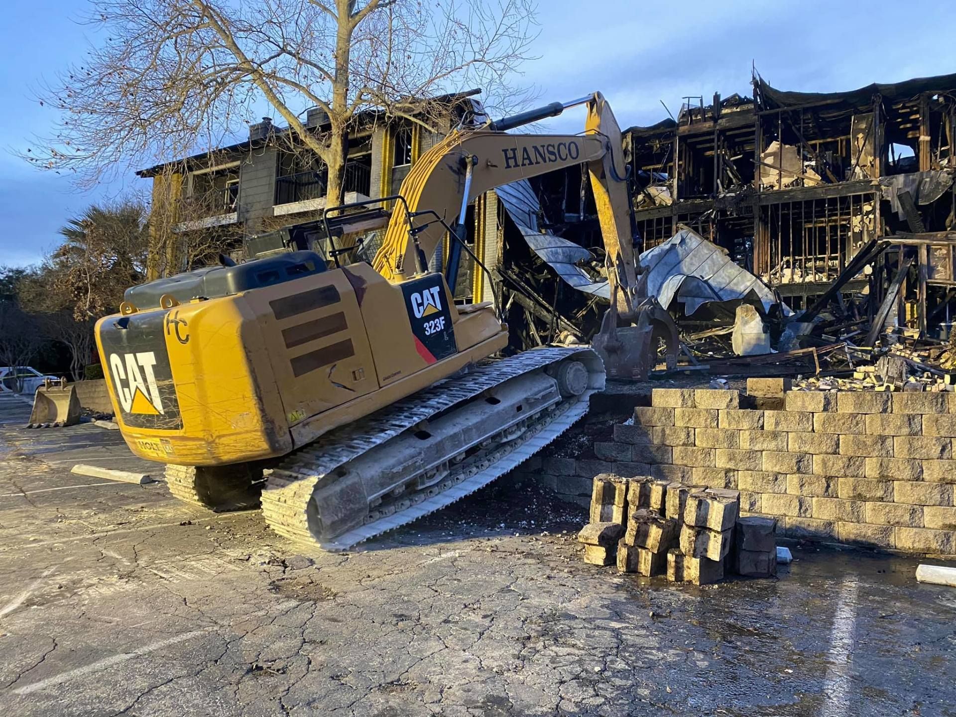 Yellow excavator demolishing a charred building, brick wall in foreground.