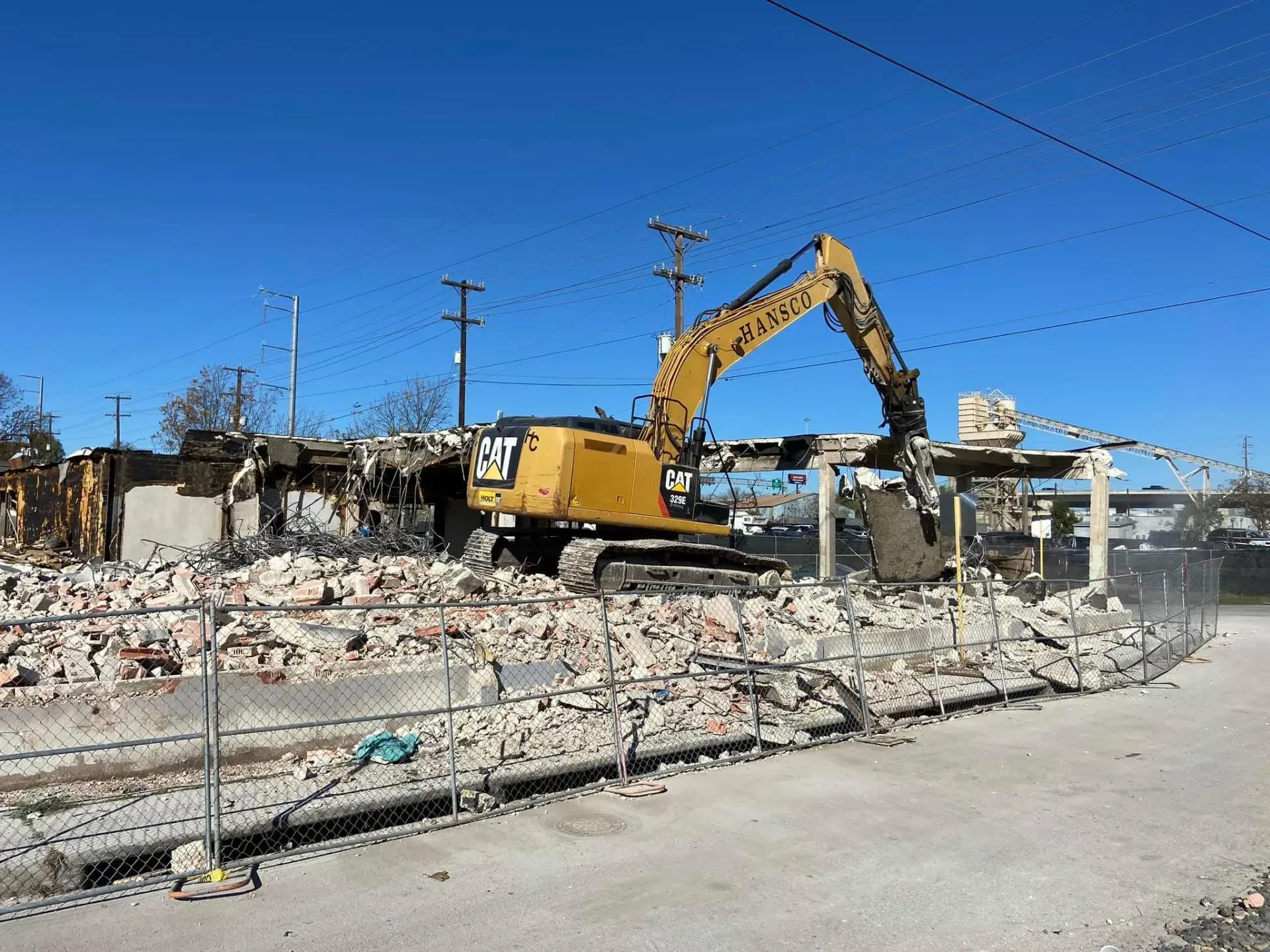 Yellow excavator demolishing a building on a sunny day. Debris and chain-link fence visible.