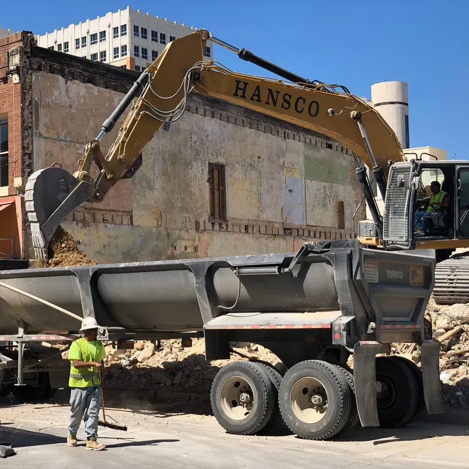 Excavator loading dirt into a dump truck during a building demolition. A worker stands nearby.