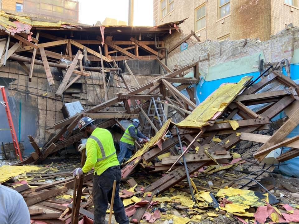 Workers in high-visibility vests demolishing a wooden structure with exposed beams; blue and yellow colors.