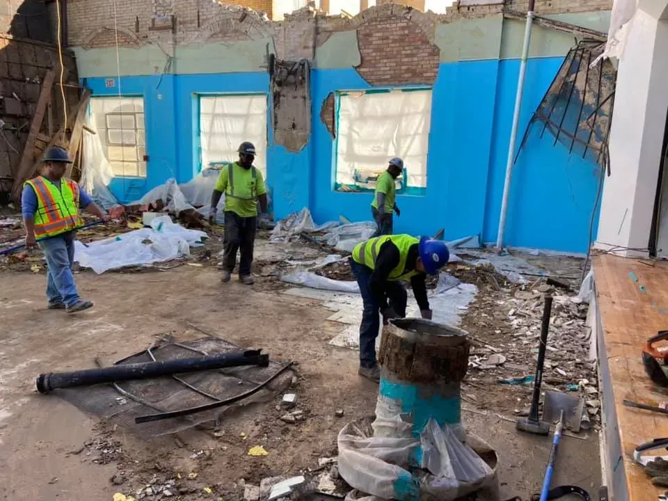 Construction workers inside a building undergoing demolition. Blue walls, debris, and tools are visible.