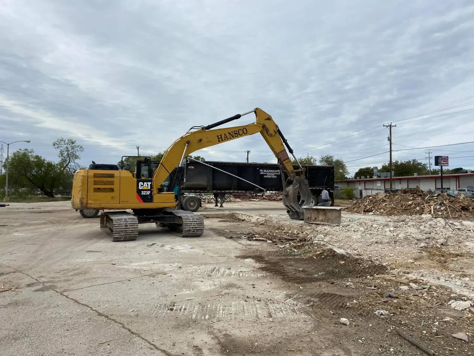 Yellow excavator loading debris into a black trailer at a demolition site under cloudy skies.