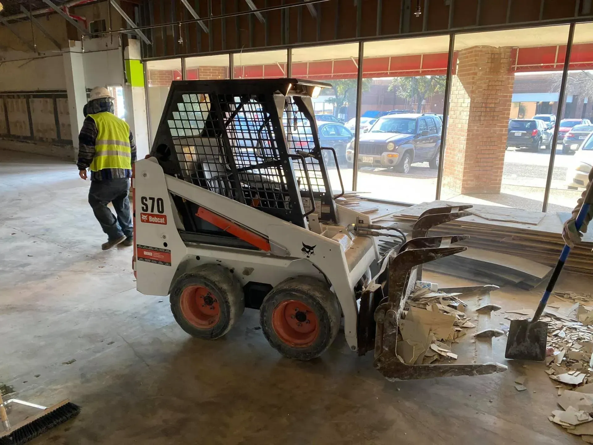 White Bobcat skid-steer loader removing debris inside a building. A worker in safety gear stands nearby.