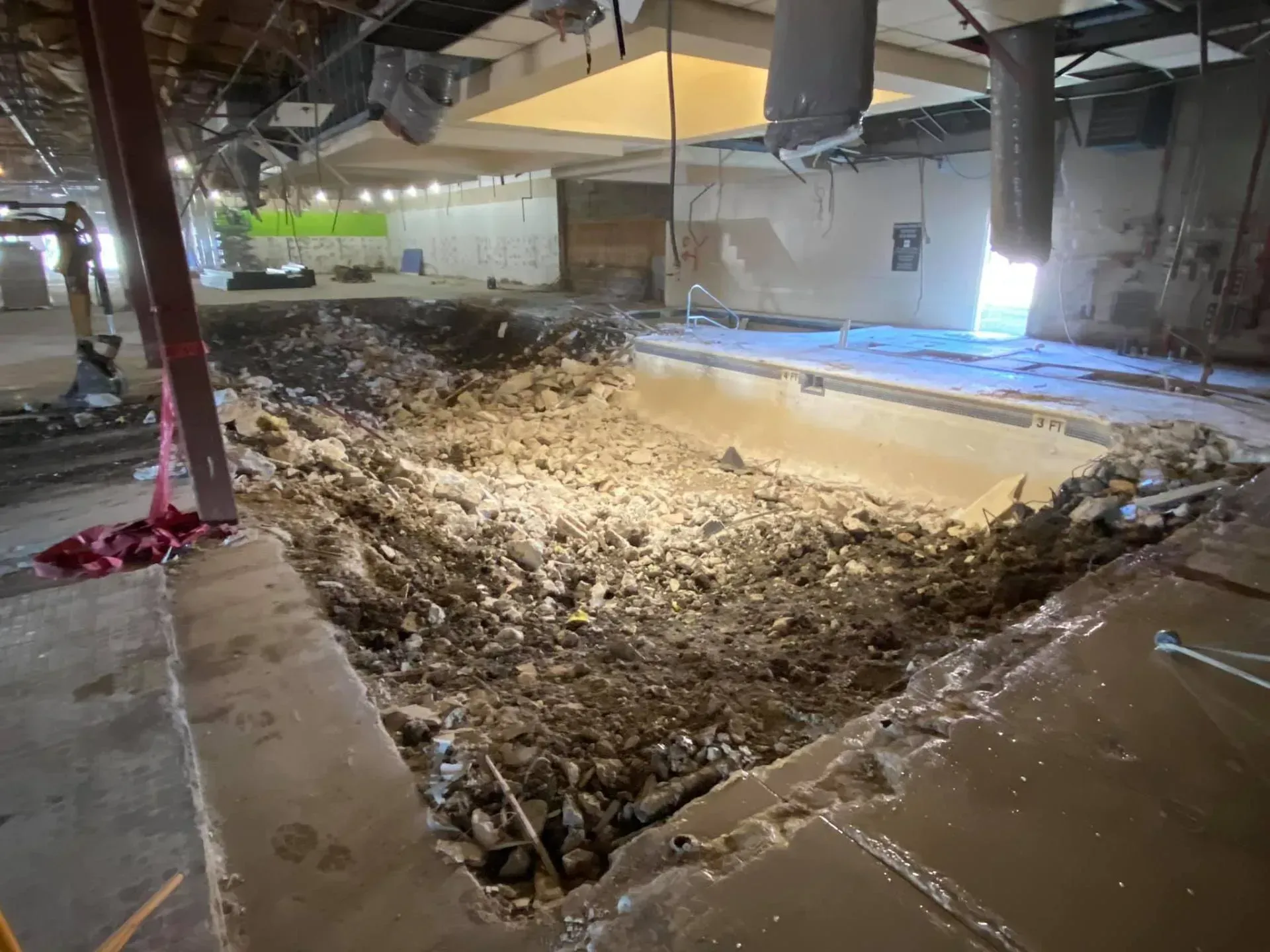 Pool demolition site. Debris fills a partially excavated pool. Gray and brown materials, open ceiling.