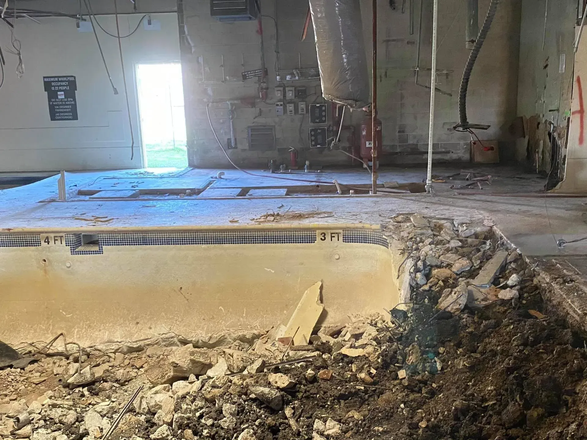 Demolished indoor pool; debris in foreground, doorway with sunlight in background.