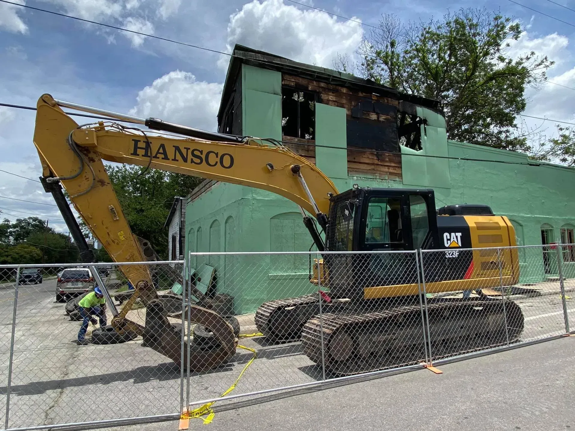 Yellow excavator demolishing a green building, fenced off, with a worker nearby.
