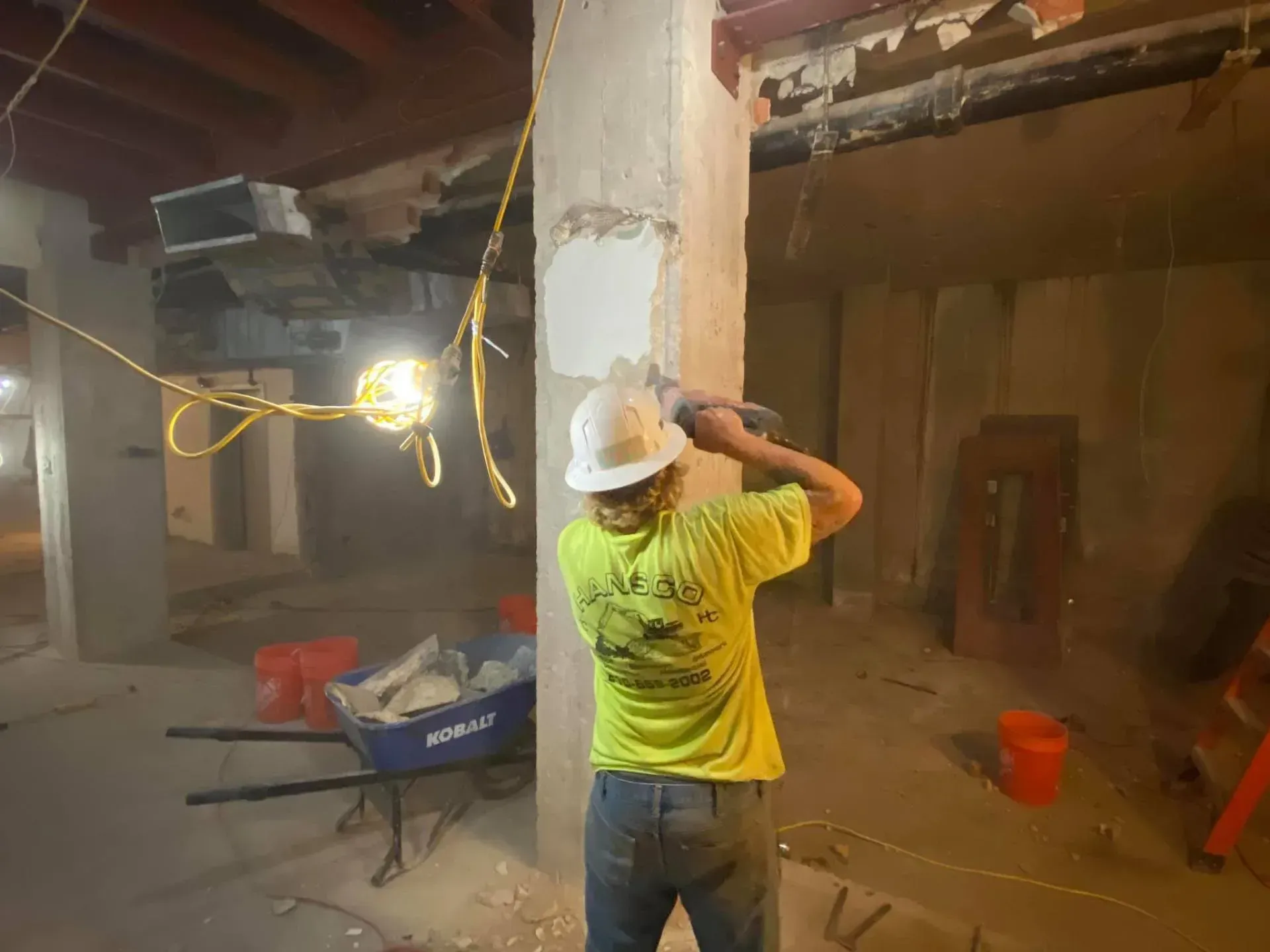 Construction worker using a power tool on a pillar in a basement. Dusty, with safety gear.