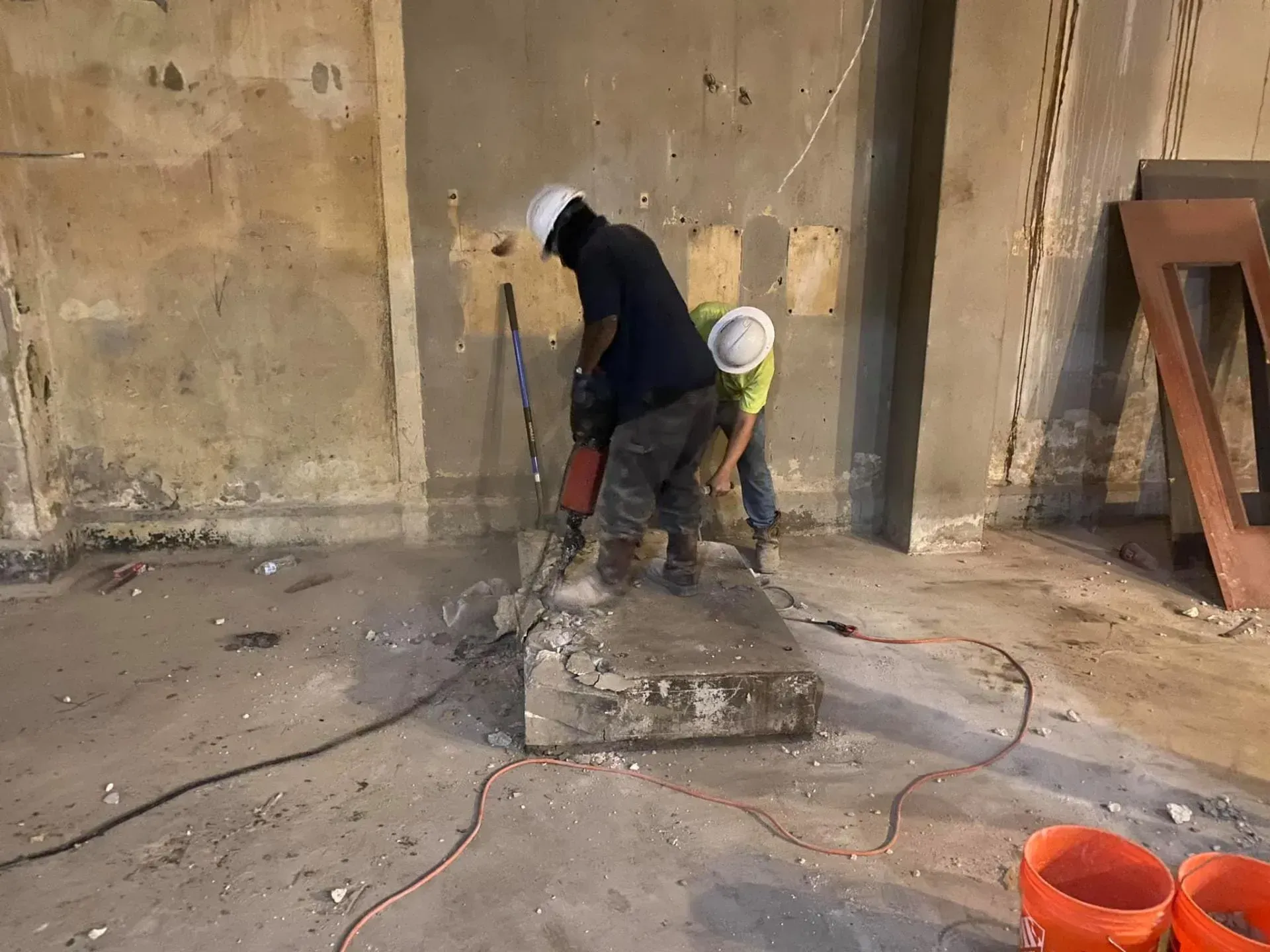 Two workers in hard hats use a jackhammer to break up a concrete block in a construction setting.