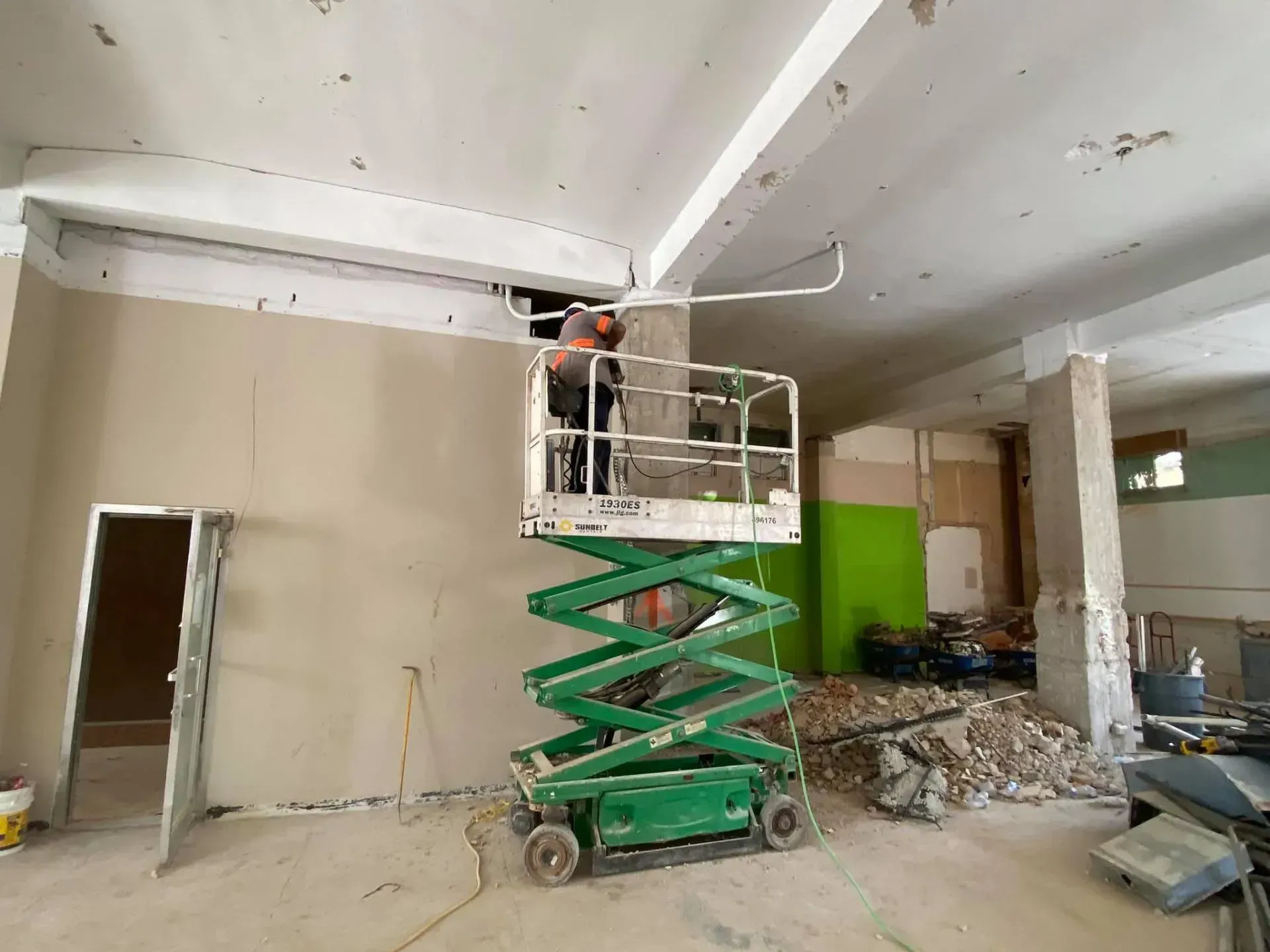Construction worker on scissor lift working on ceiling in a room with exposed beams and debris.