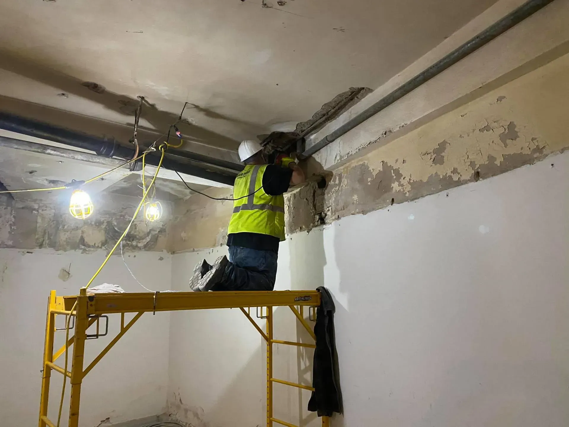 Worker on scaffolding drills into ceiling, wearing safety gear in a room with exposed pipes and damaged walls.
