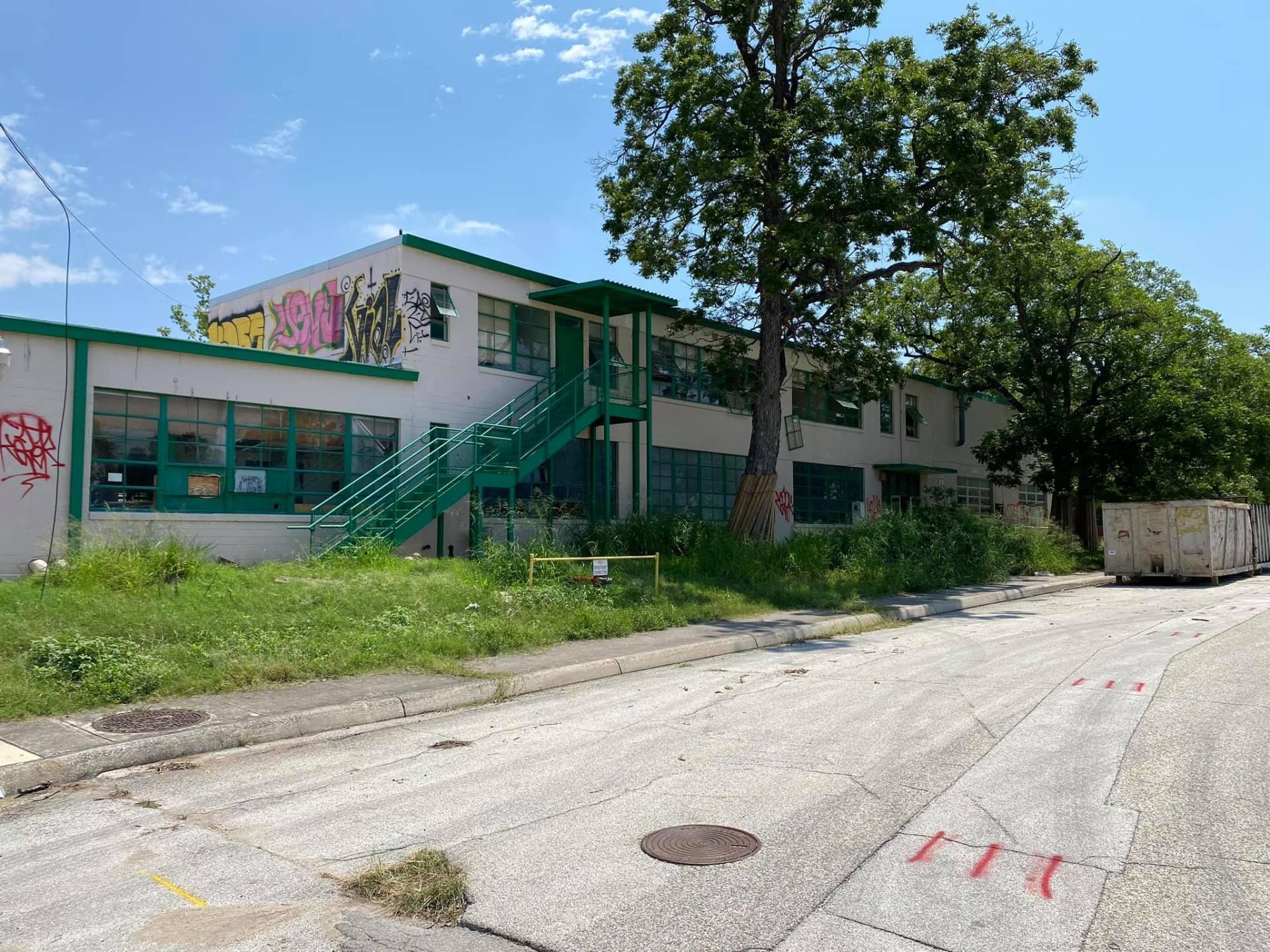 Two-story, white building with green trim and stairs, overgrown grass, and graffiti. Tree in front.
