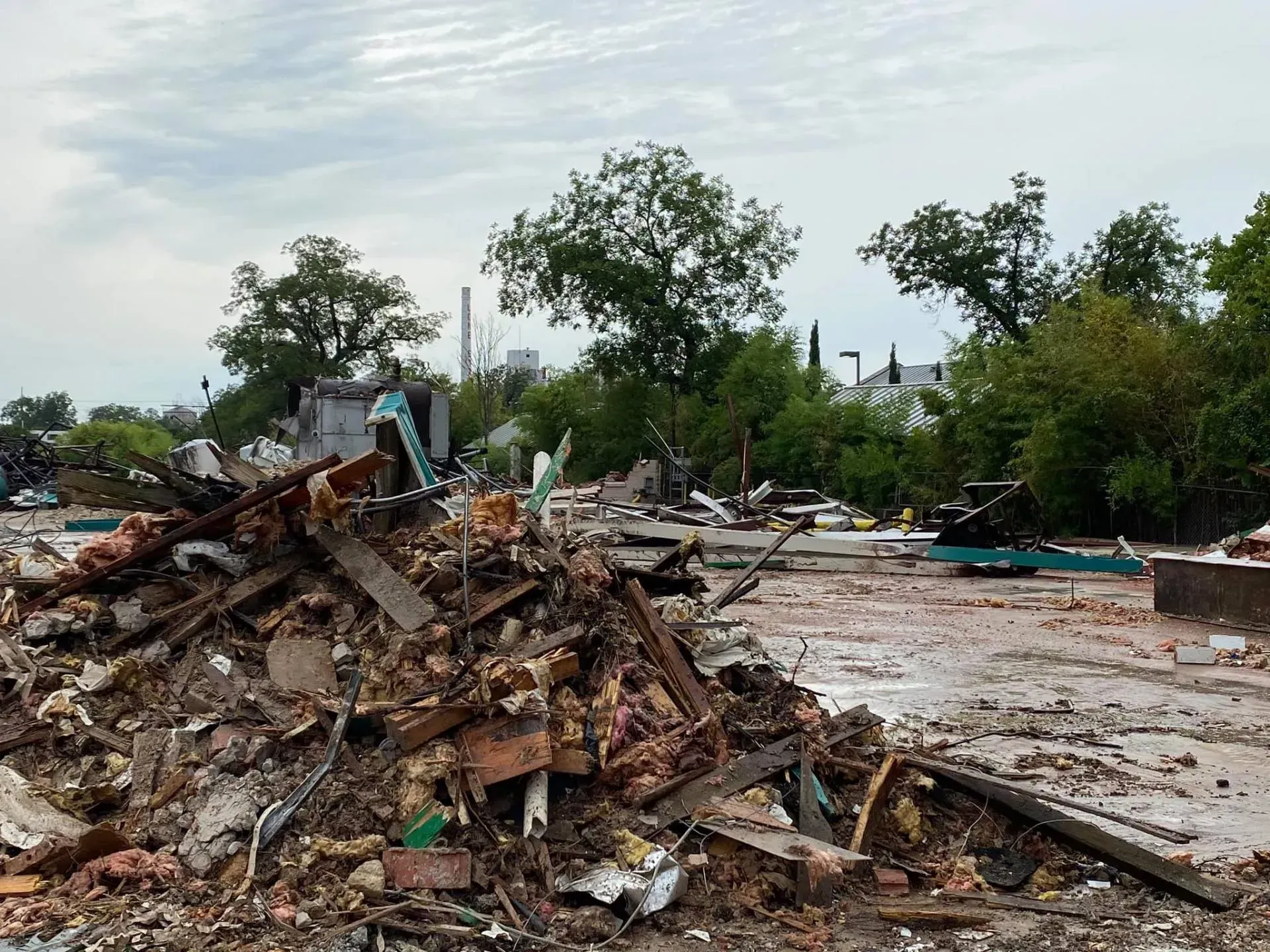 Debris pile and cleared concrete lot, trees in background under overcast sky.