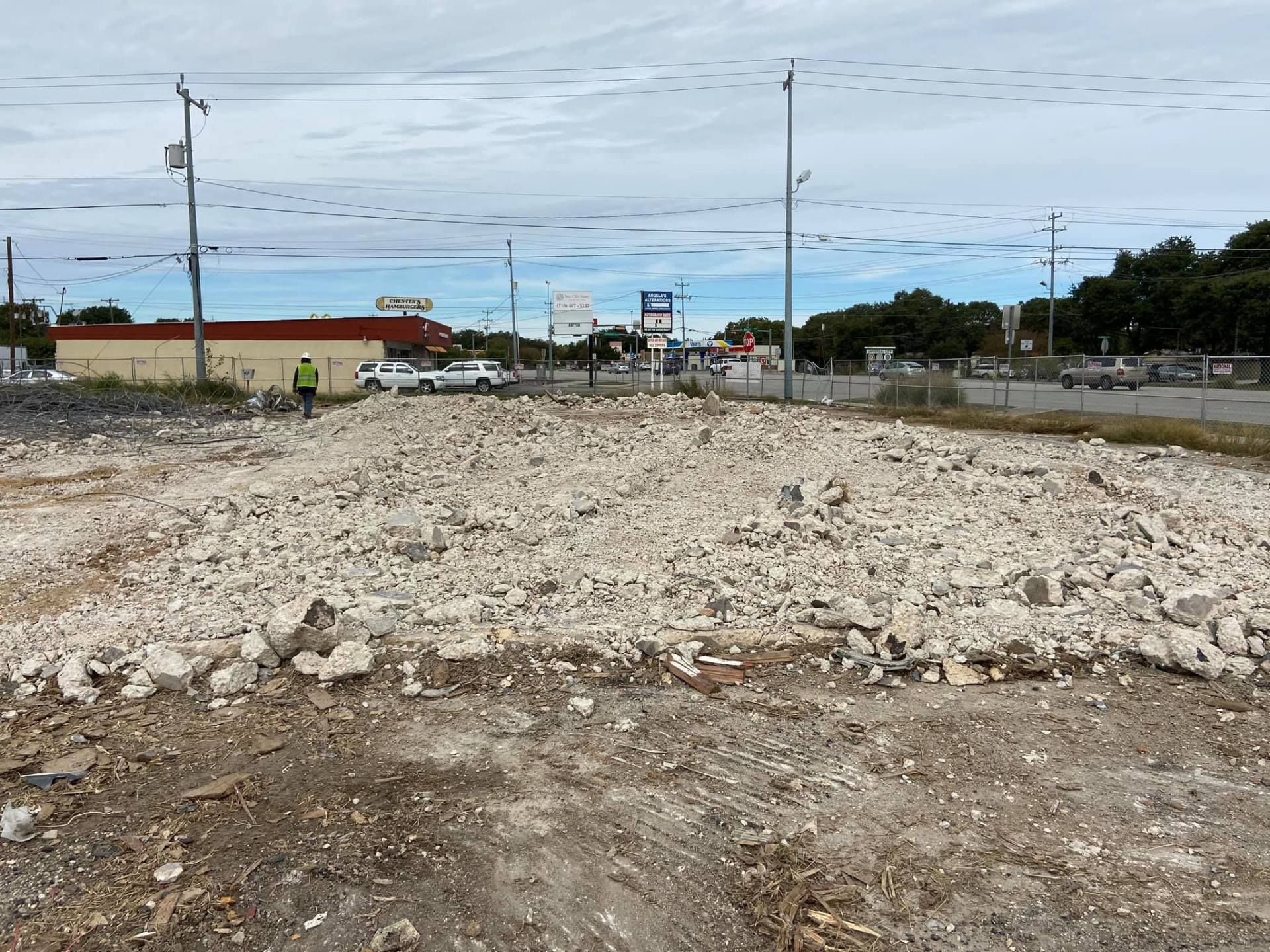 A construction site with rubble in the foreground, with buildings, cars, and a worker in the background under a cloudy sky.