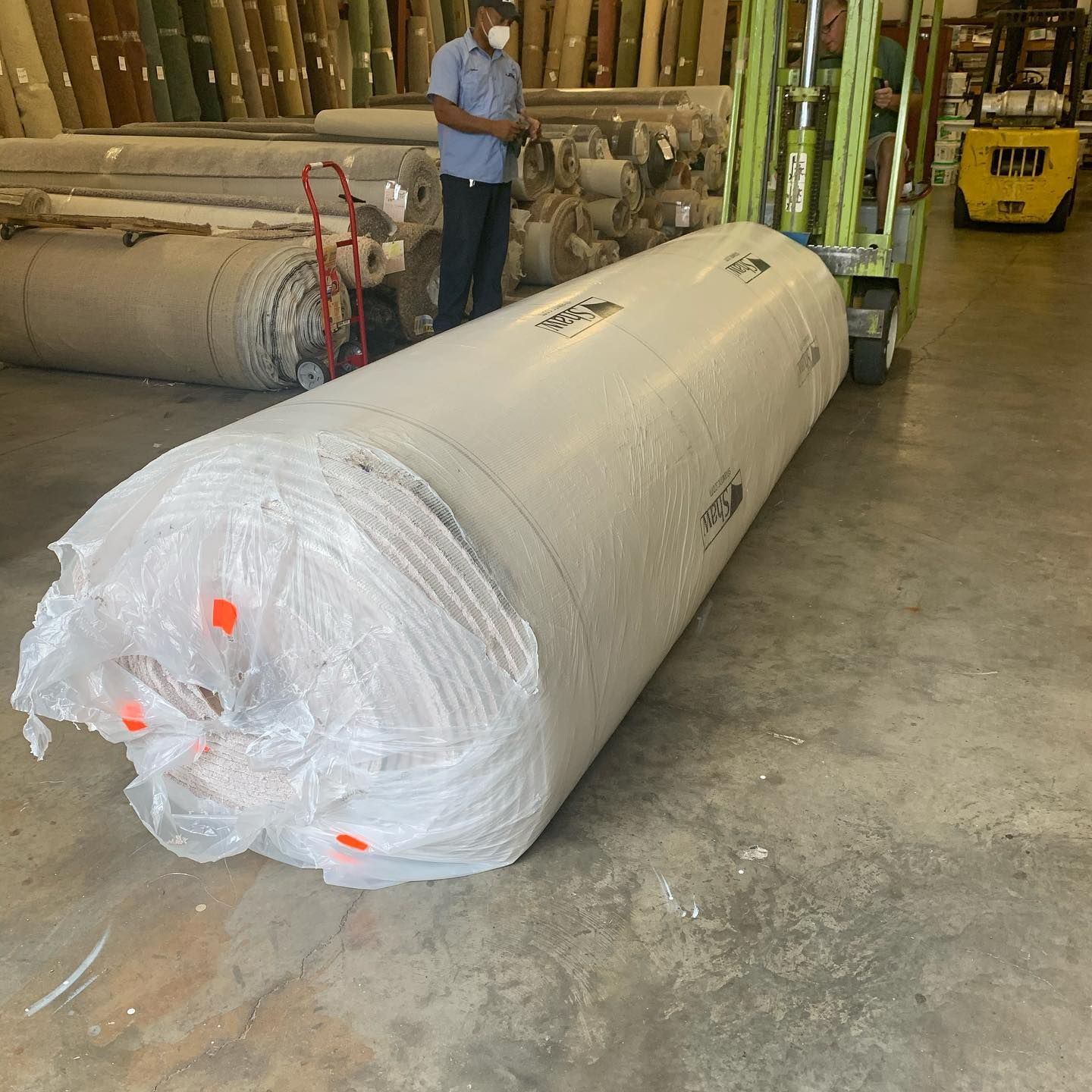 Large carpet roll wrapped in plastic being moved by a forklift in a warehouse.