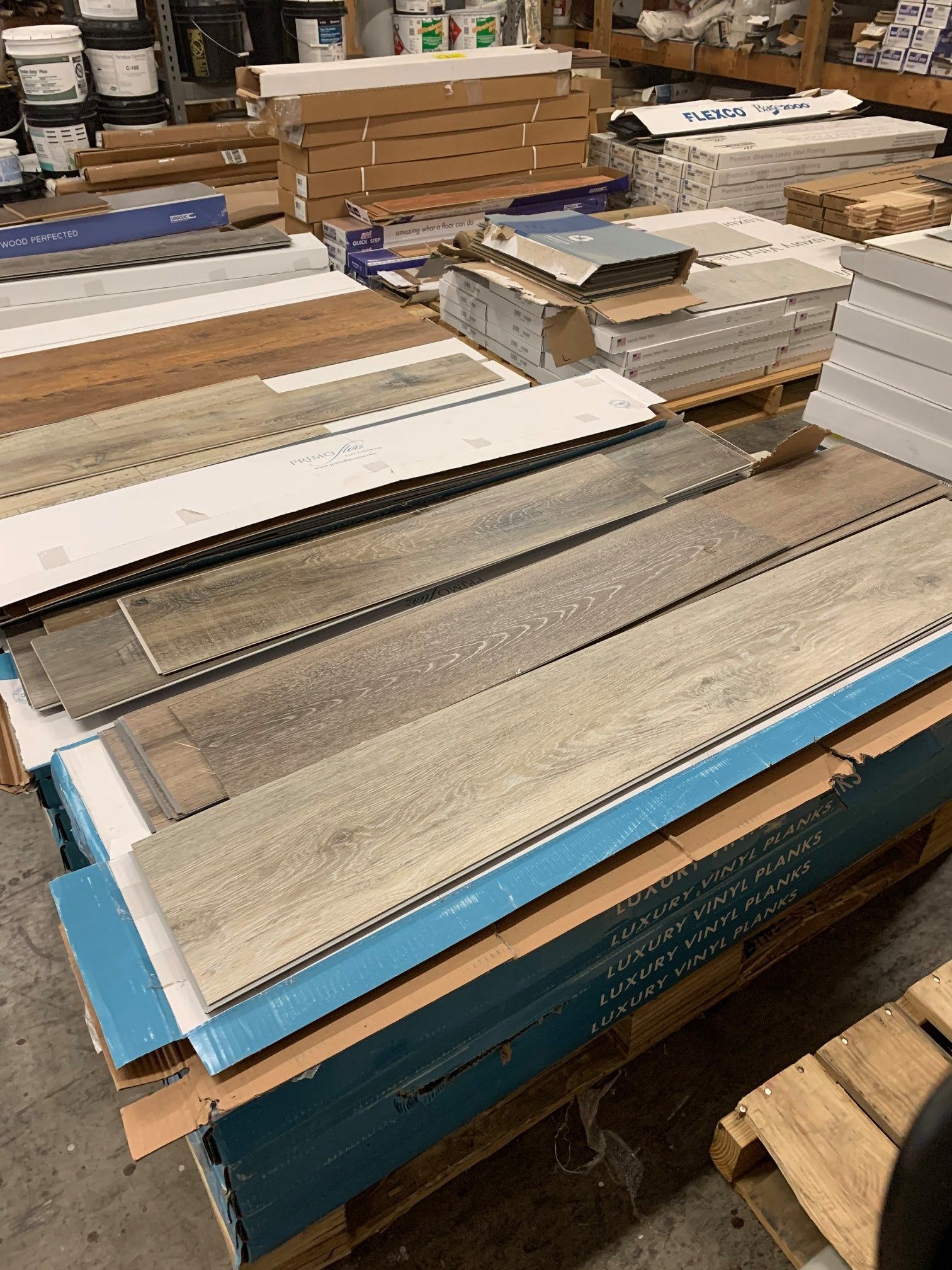Stacks of wood flooring samples in a warehouse. Various wood tones, stored on pallets, boxes visible.