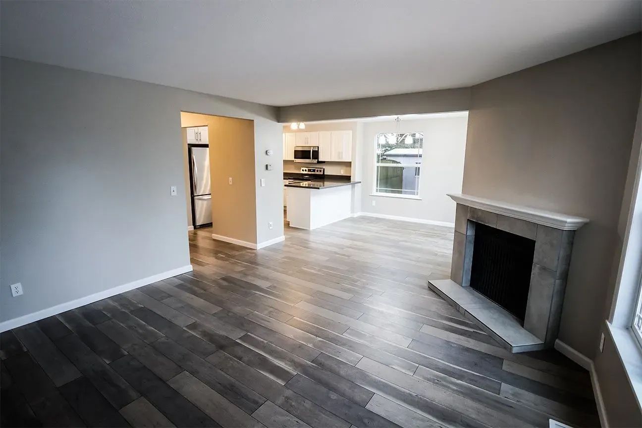Empty living room with gray walls, dark wood floors, and a fireplace opening to a bright kitchen.