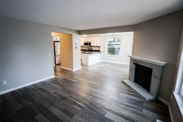 Empty living room with gray walls, dark wood floors, and a fireplace opening to a bright kitchen.