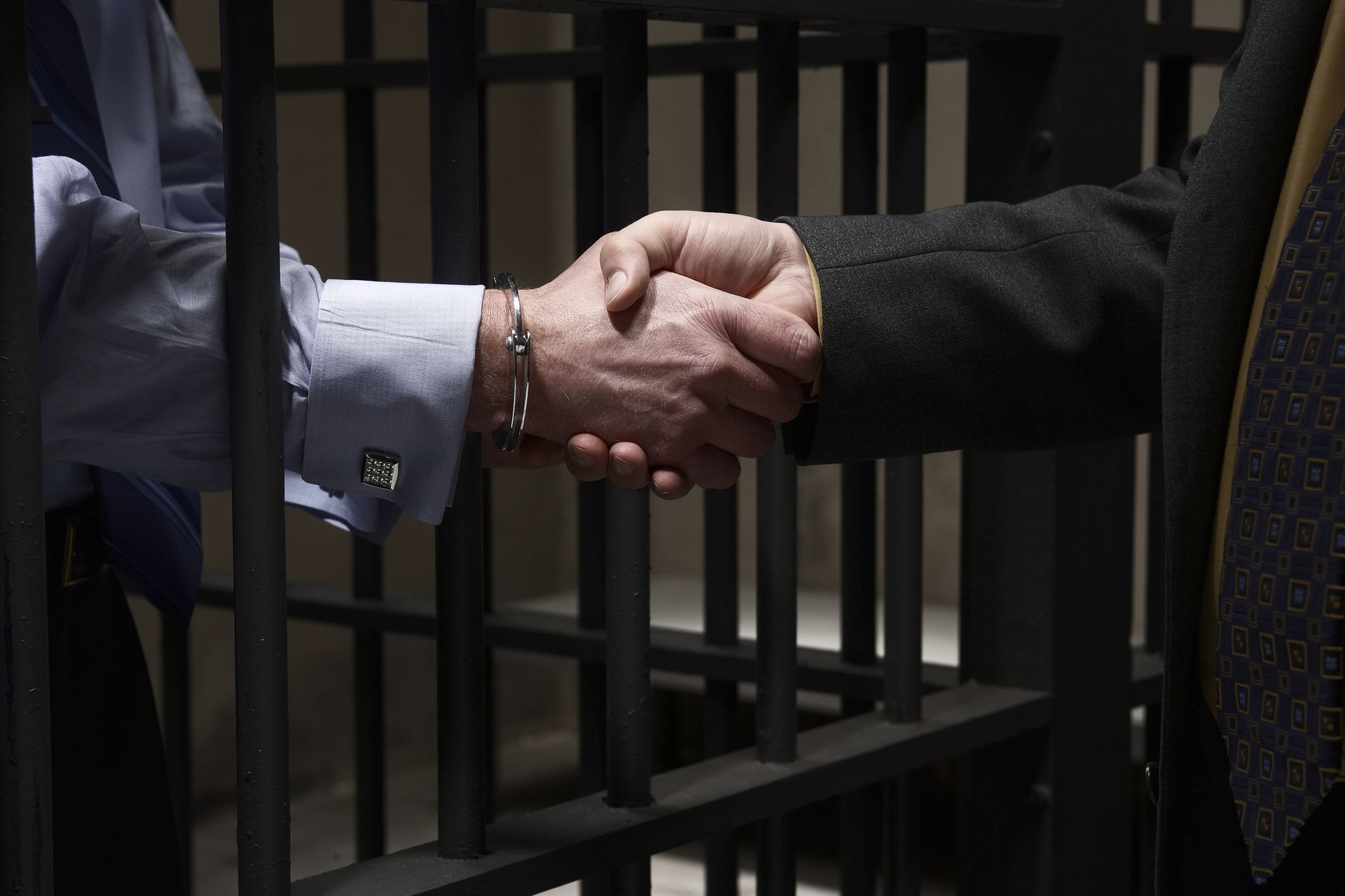 Handshake through prison bars. Two men's arms meeting. One in a suit, the other in a button-down shirt.