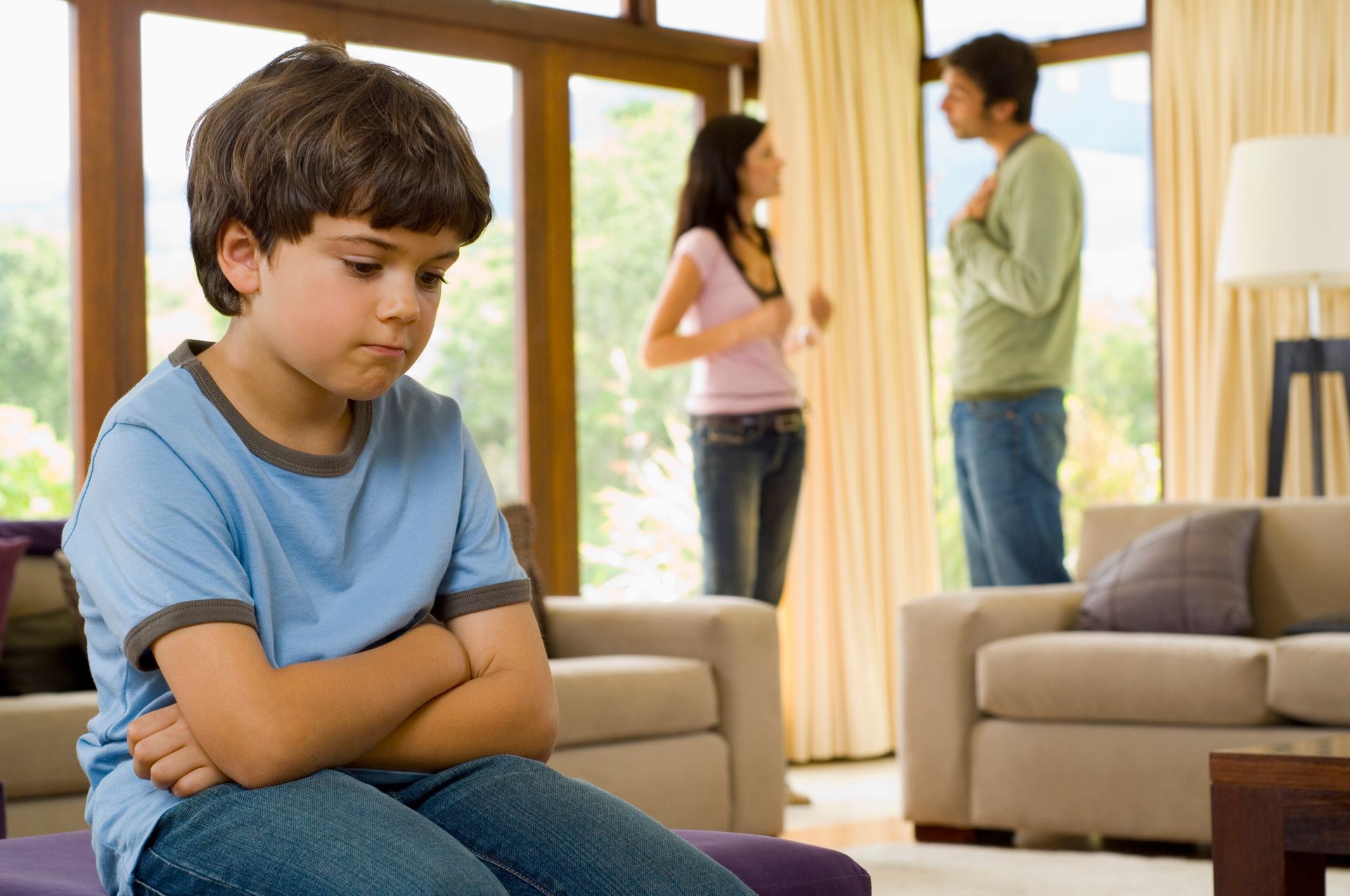 Boy with arms crossed looks sad while parents argue in the background of a living room.