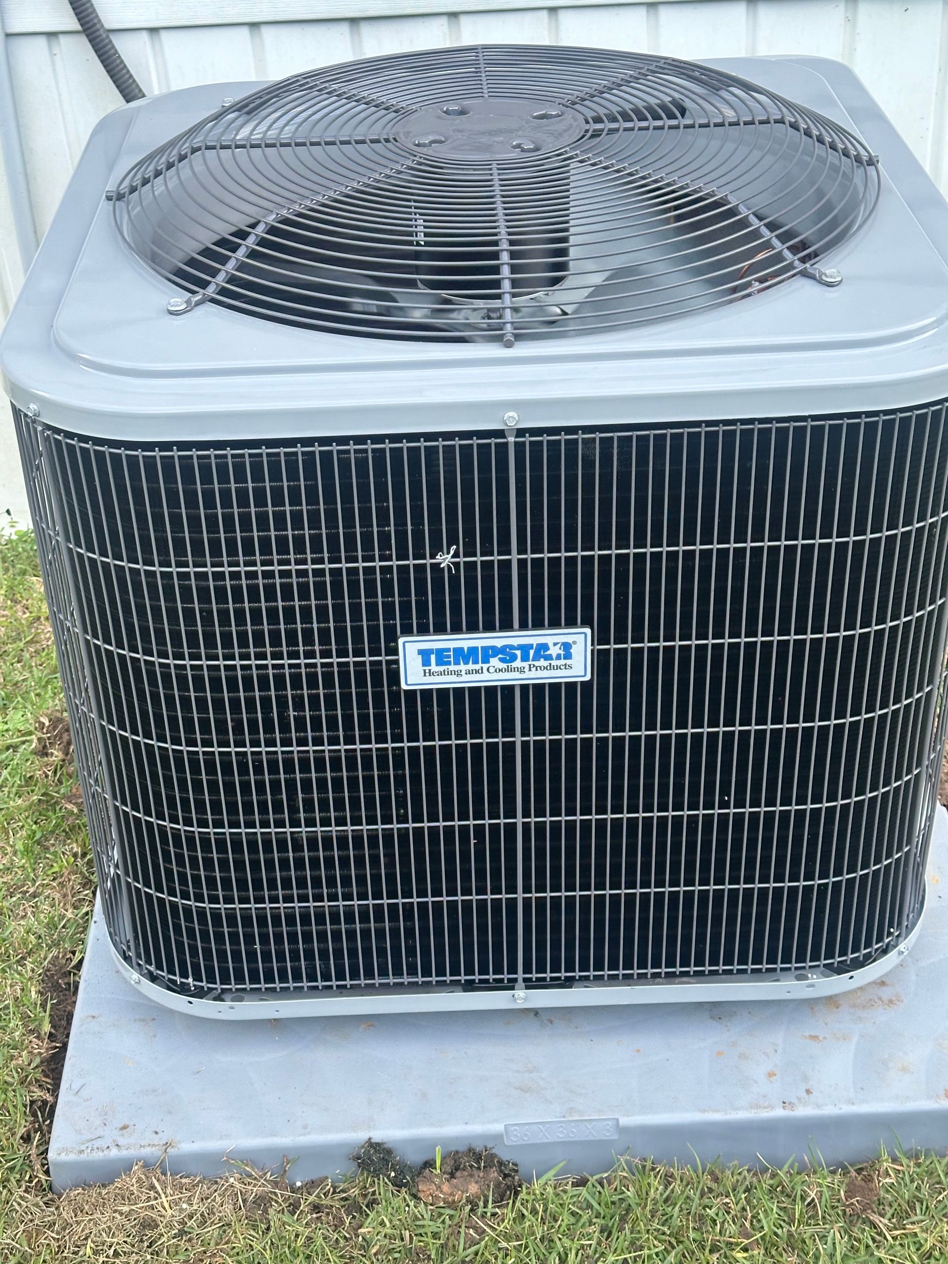 A tempest air conditioner is sitting in the grass outside of a house.