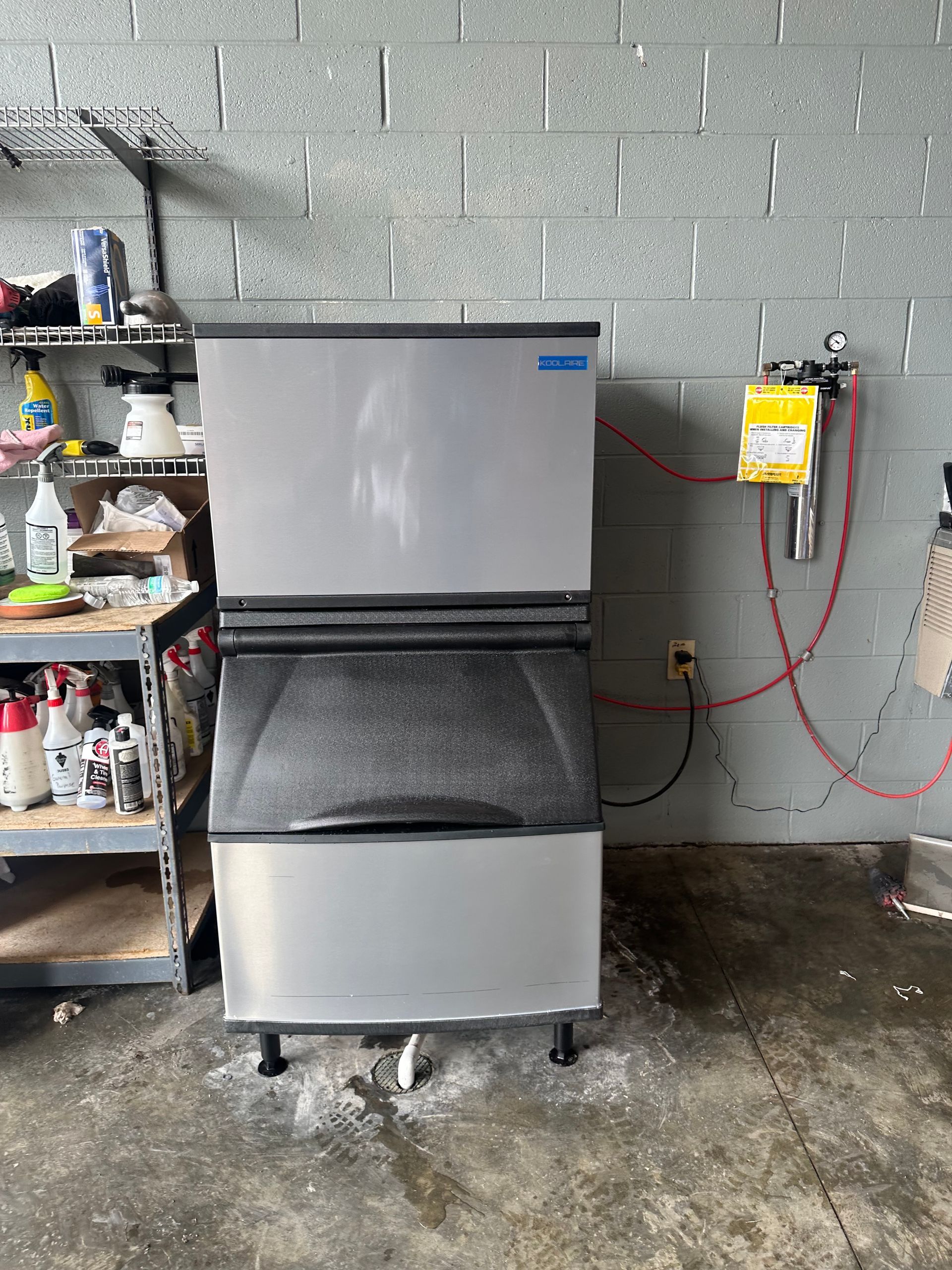 A stainless steel ice machine is sitting in a garage next to a brick wall.