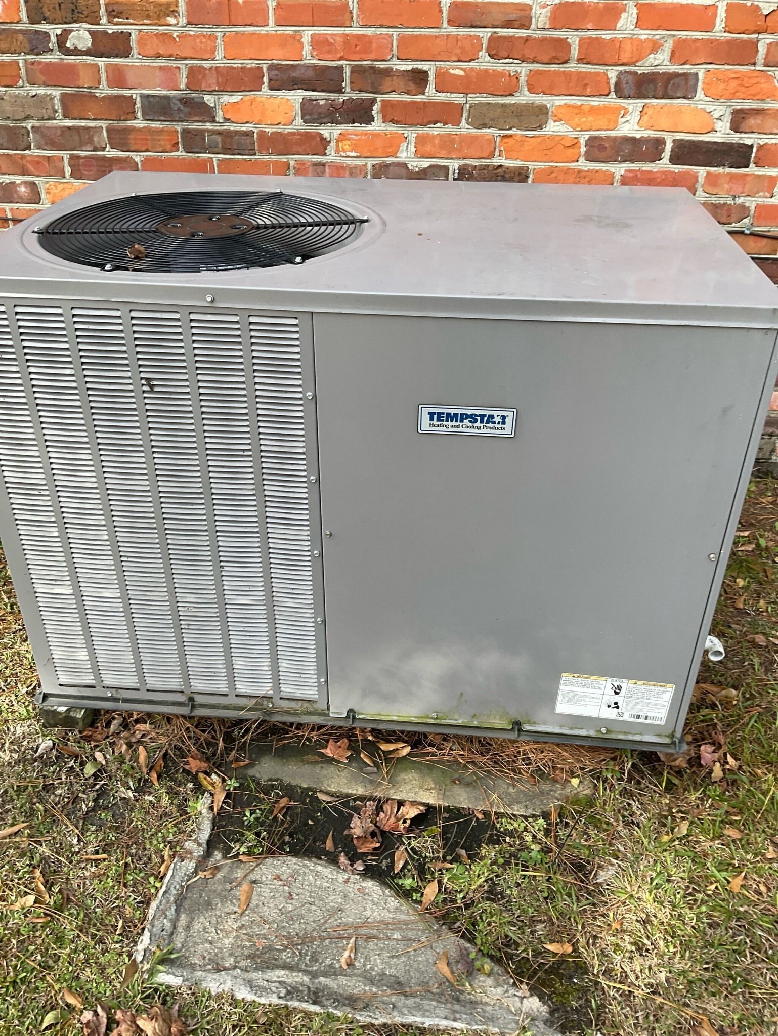 A gray air conditioner is sitting on the ground in front of a brick wall