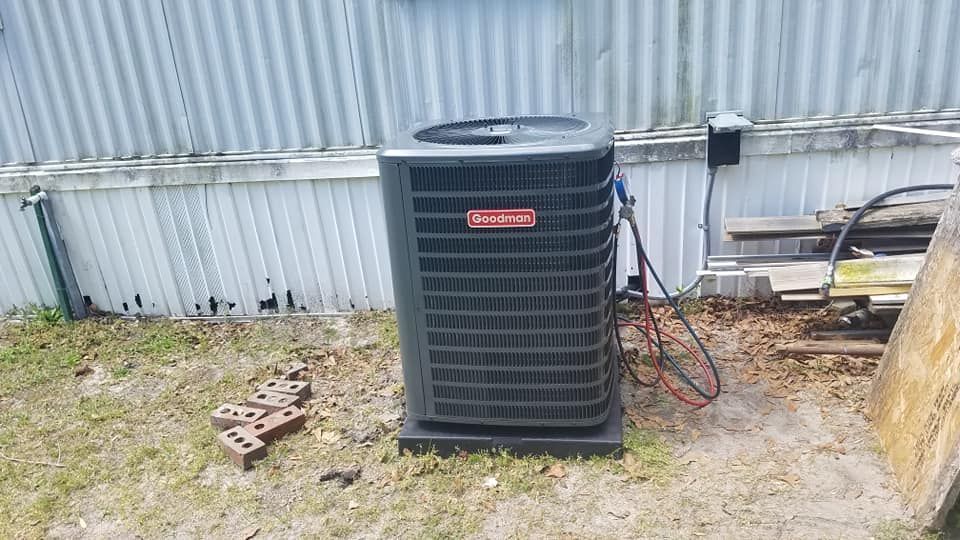 A black air conditioner is sitting in the grass in front of a white fence