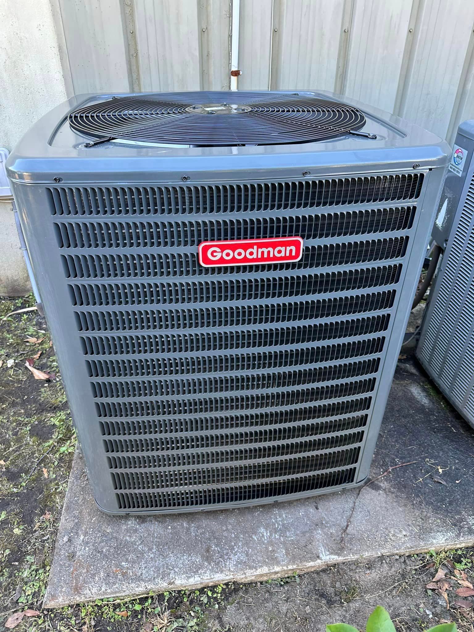 A goodman air conditioner is sitting on the ground next to a building.