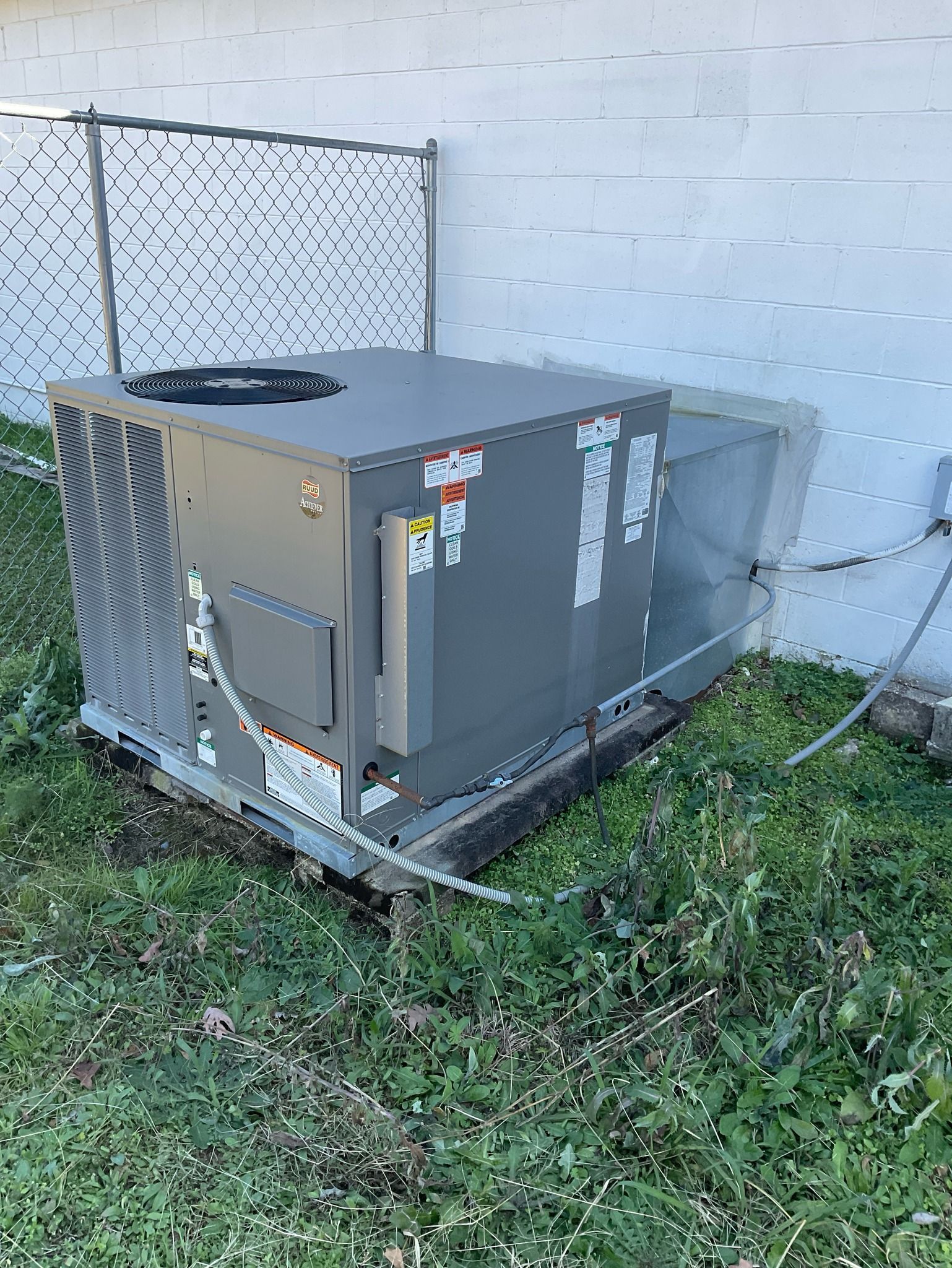 A large air conditioner is sitting in the grass next to a chain link fence.